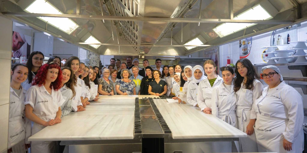 Profesyonel bir mutfakta, beyaz önlükler giymiş kalabalık bir grup kursiyer ve eğitmen, uzun mermer bir çalışma tezgahının iki yanında yan yana dizilmiş şekilde kameraya gülümsüyor. Çoğunluğu kadınlardan oluşan grubun önündeki tezgahın üzerinde bazı belgeler ve küçük objeler bulunuyor. Arka planda paslanmaz çelik mutfak ekipmanları, davlumbazlar ve duvarda pastacılıkla ilgili görseller yer alıyor.