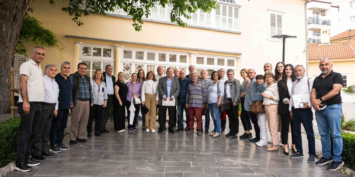 Tarihi bir binanın önündeki taş döşeli avluda yan yana dizilmiş, farklı yaş gruplarından oluşan kalabalık bir grup insan, kameraya bakarak poz veriyor. Gruptaki kadın ve erkekler günlük kıyafetler içinde, rahat bir duruş sergiliyor. Arka planda beyaz pencereli, klasik mimariye sahip bir yapı ve ağaç gölgesi görülüyor.