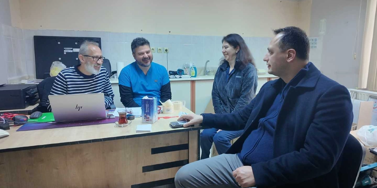 Ofis ortamında bir masa etrafında toplanmış dört kişi sohbet ediyor. Masanın sol tarafında oturan, çizgili kazaklı ve gözlüklü bir adamın önünde açık bir dizüstü bilgisayar bulunuyor. Yanında mavi forma giymiş bir erkek, karşısında ise koyu renkli bir ceket giymiş başka bir erkek ve yanında duran bir kadın yer alıyor. Masanın üzerinde bir bardak çay ve çeşitli ofis malzemeleri bulunuyor.