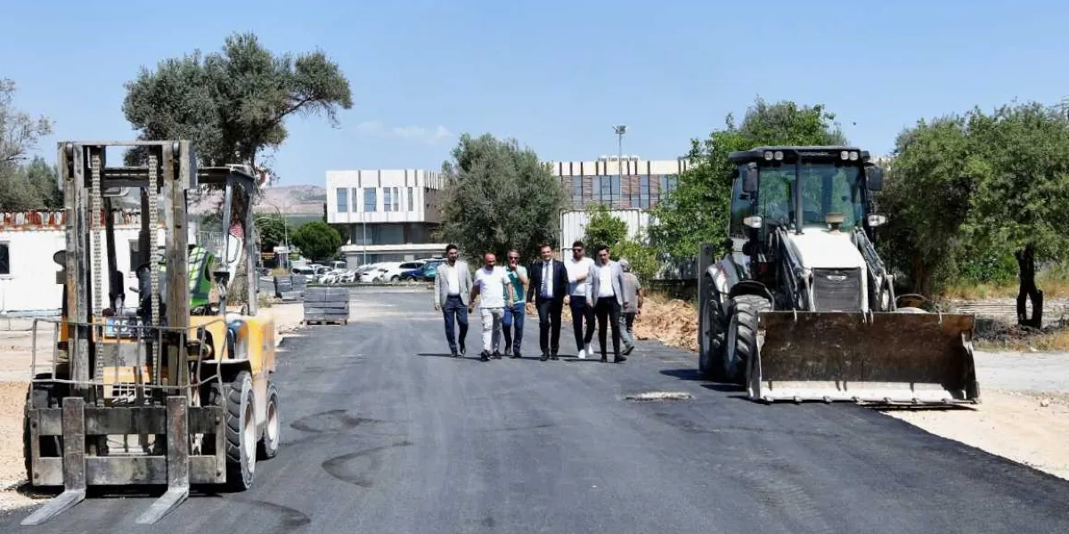 Yeni asfaltlanmış bir yol üzerinde yürüyen, aralarında Bornova Belediye Başkanı'nın da bulunduğu bir grup yetkili. Yolun sol tarafında sarı renkli bir forklift, sağ tarafında ise büyük bir iş makinesi park halinde duruyor. Arka planda ağaçlar ve modern bir bina yapısı görülüyor.