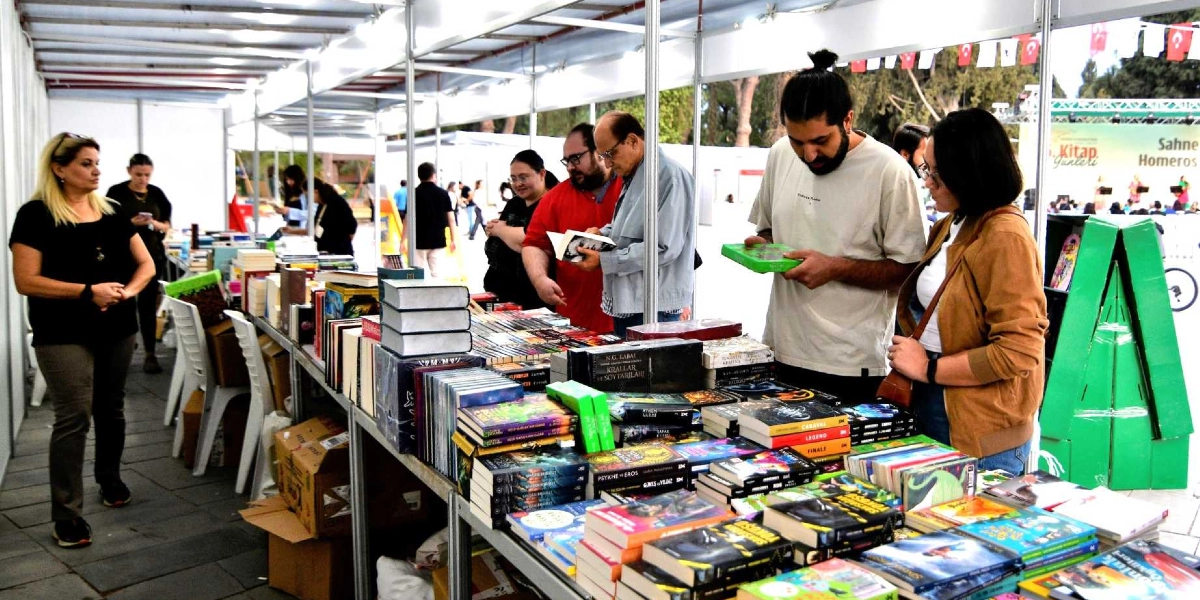 Bornova'daki bir kitap fuarında, üstü kapalı stantların altında yan yana dizilmiş masaların üzerinde sergilenen kitapları inceleyen ziyaretçiler. Ön planda, elinde yeşil kapaklı bir kitap tutan uzun saçlı bir genç adam ve yanında duran gözlüklü bir kadın kitaplara bakıyor. Arka planda ise stantlar arasında dolaşan diğer ziyaretçiler ve kitap stantlarının genel görünümü yer alıyor. Sağ tarafta üzerinde 