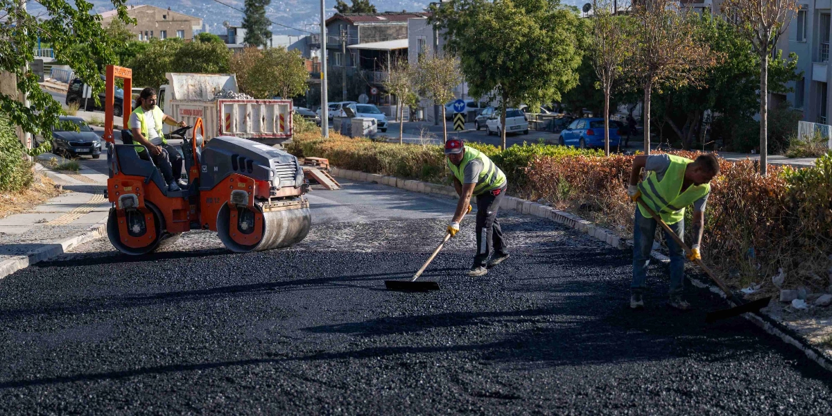 Bornova Belediyesi ekipleri, bir sokakta asfalt serim ve düzeltme çalışması yapıyor. Sarı ikaz yelekli bir operatör silindir aracını kullanırken, diğer iki işçi ellerindeki tırmıklarla yeni dökülen asfaltı düzeltiyor. Arka planda park halindeki araçlar ve ağaçlı bir yol kenarı görülüyor.