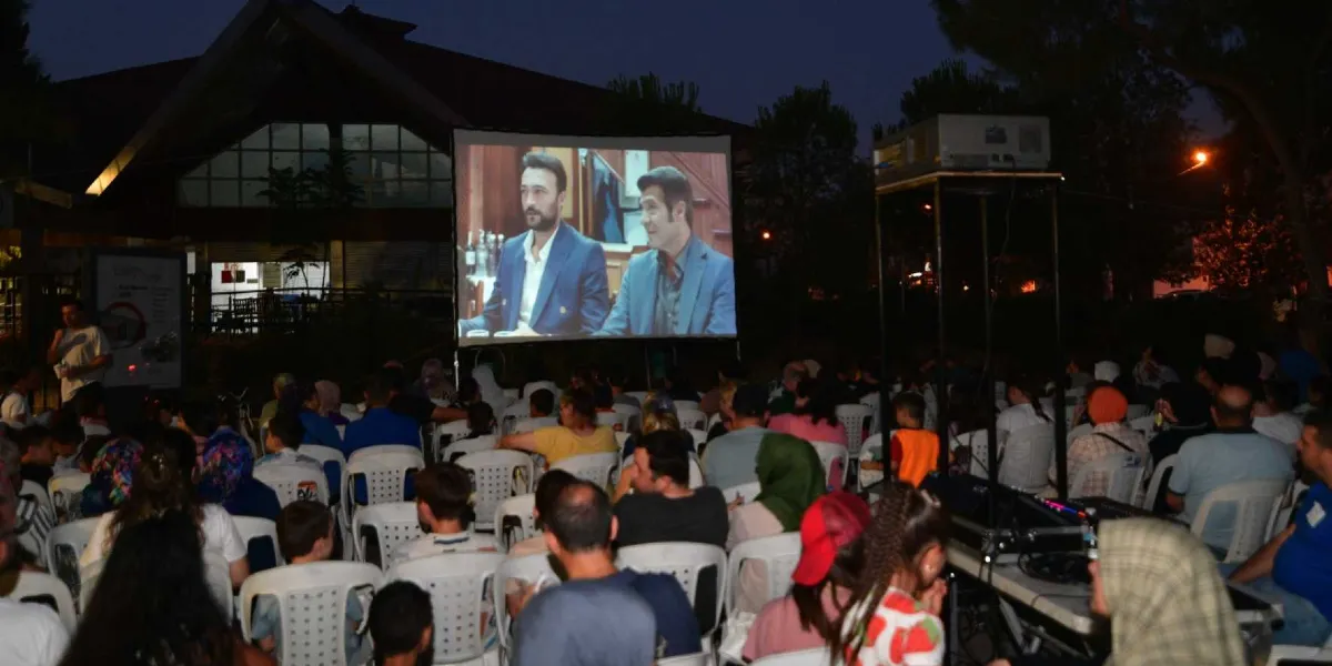 Akşam saatlerinde açık havada kurulan dev bir perdede film izleyen kalabalık bir izleyici grubu. Beyaz plastik sandalyelere oturan izleyiciler, perdede görünen iki erkek oyuncunun yer aldığı sahneyi seyrediyor. Arka planda ağaçlar ve bir bina silüeti yer alırken, sağ tarafta yüksek bir stand üzerinde projeksiyon cihazı bulunuyor.