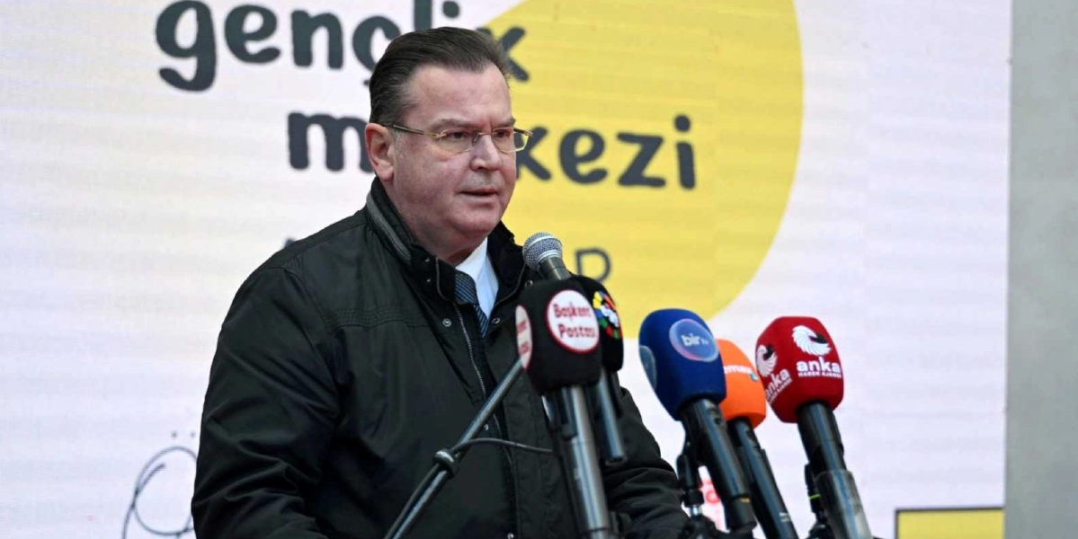 Gözlüklü, koyu renk ceketli bir yetkili, arkasında 
