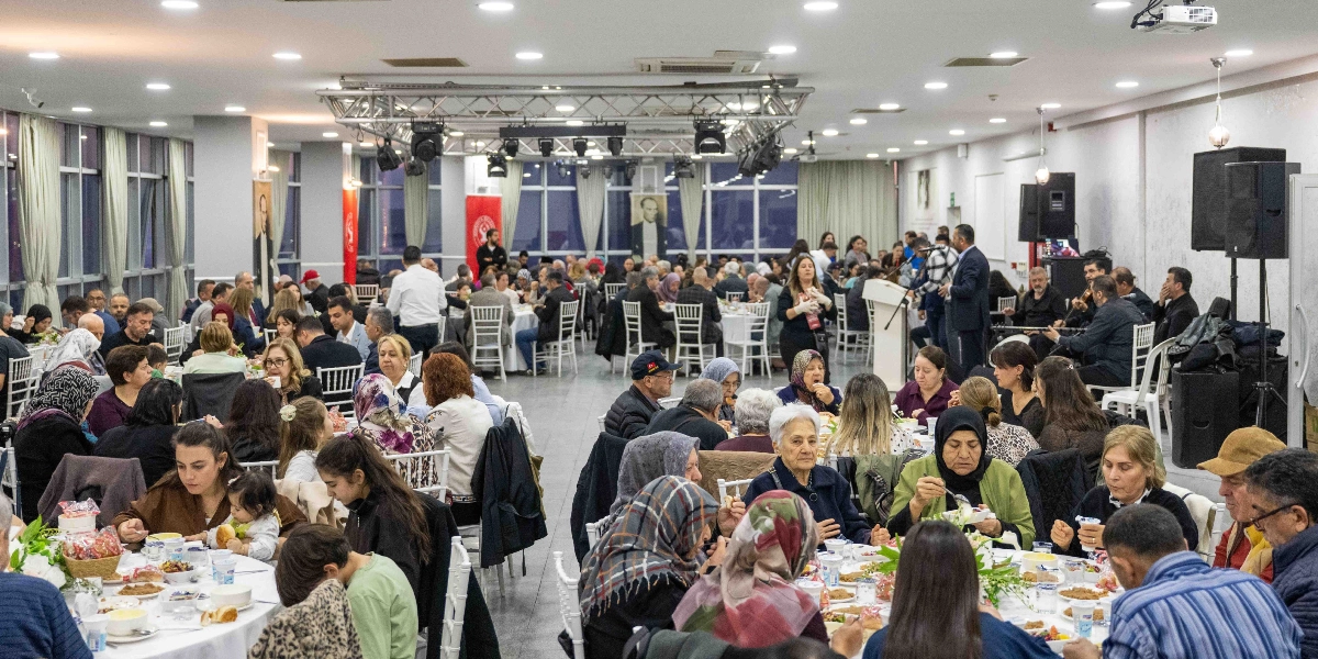 Geniş bir etkinlik salonunda, çok sayıda yuvarlak masanın etrafında toplanmış kalabalık bir grup insan yemek yiyor. Salonun arka kısmında bir kürsü ve müzisyenler bulunurken, duvarlarda Atatürk portreleri asılı. Katılımcılar arasında her yaştan kadın ve erkek yer alıyor; masalarda yemek tabakları, içecekler ve ekmek sepetleri bulunuyor. Ortam, toplu bir yemek daveti veya iftar programı havasında geçiyor.