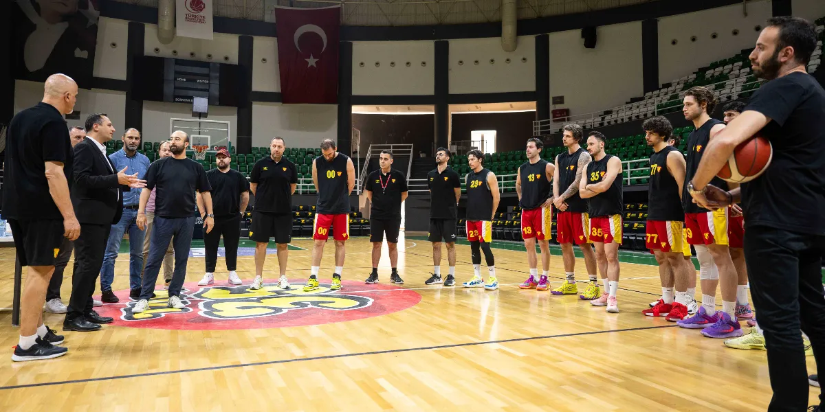 Bornova Belediye Başkanı Ömer Eşki, bir basketbol sahasında Göztepe Basketbol takımı oyuncuları ve teknik ekibiyle bir araya gelerek sohbet ediyor. Oyuncular siyah formaları ve sarı-kırmızı şortlarıyla sahada yan yana dizilmiş şekilde dururken, teknik ekip ve Başkan Eşki sol tarafta yer alıyor. Arka planda Türk bayrağı ve basketbol sahası tribünleri görülüyor.