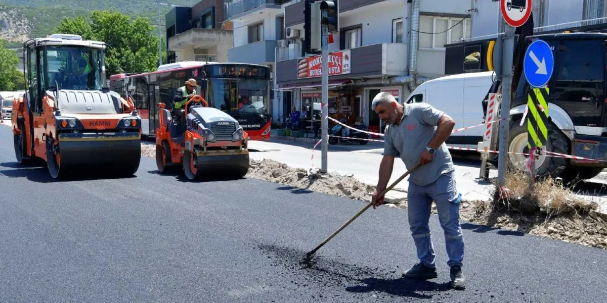 Belediye ekiplerinin bir şehir caddesinde yürüttüğü asfalt yenileme çalışması sırasında, bir işçi elindeki tırmıkla yeni dökülen asfaltı düzeltiyor. Arka planda iki adet Hamm marka silindir aracı, bir belediye otobüsü ve yol kenarında dükkanlar yer alıyor.