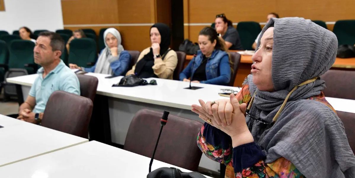 Bir toplantı salonunda, ön planda konuşma yapan başörtülü bir kadın ve onu dinleyen katılımcılar yer alıyor. Katılımcılar beyaz masaların etrafında oturmuş, dikkatle dinlerken görülüyor. Ön plandaki kadın, ellerini birleştirmiş bir şekilde konuşurken, arka planda ise farklı yaş ve giyim tarzlarından kadın ve erkeklerden oluşan bir grup dinleyici bulunuyor.