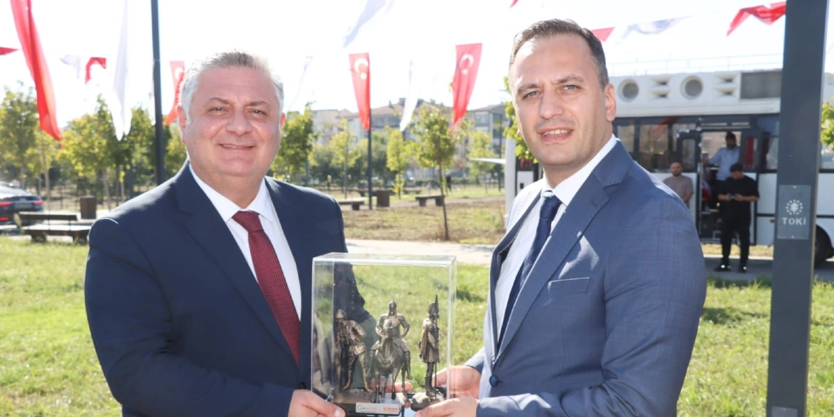 Takım elbiseli ve kravatlı iki erkek, açık havada, ellerinde içerisinde tarihi figürlerin bulunduğu cam bir plaketi birlikte tutarak gülümseyerek poz veriyor. Arka planda Türk bayrakları, ağaçlar ve üzerinde TOKİ yazısı bulunan bir araç yer alıyor.