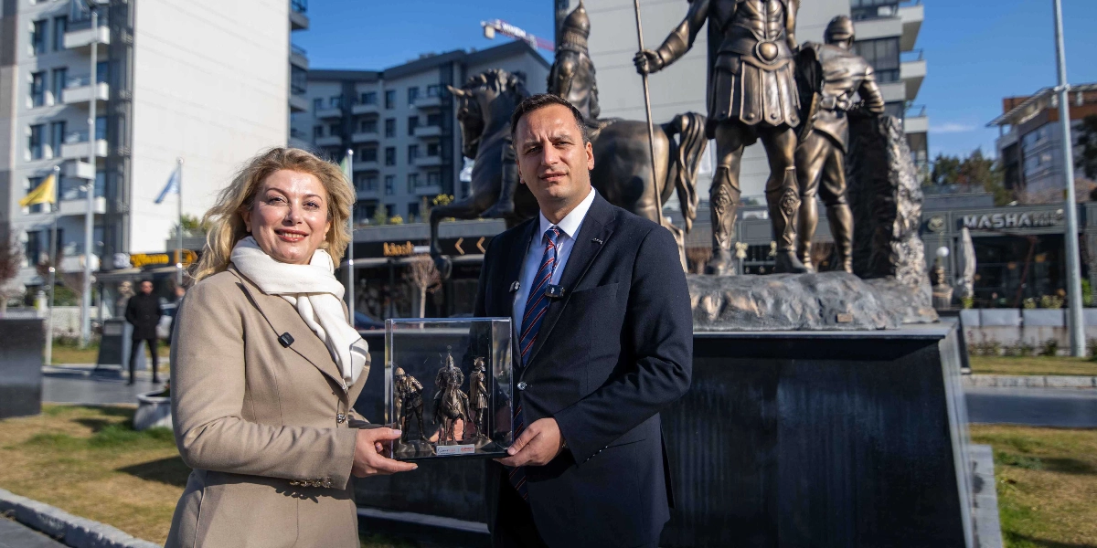 CHP Kadın Kolları Başkanı Asu Kaya ve Bornova Belediye Başkanı Ömer Eşki, açık havada, arkalarında büyük bir bronz heykel grubuyla yan yana duruyor. Asu Kaya bej rengi bir ceket ve beyaz bir atkı giymiş, Ömer Eşki ise lacivert bir takım elbise ve kravat takmış durumda. İkili, ellerinde şeffaf bir kutu içerisinde bulunan minyatür bir heykel figürünü birlikte tutuyor. Arka planda modern apartmanlar ve çevre düzenlemesi yapılmış bir alan görülüyor.