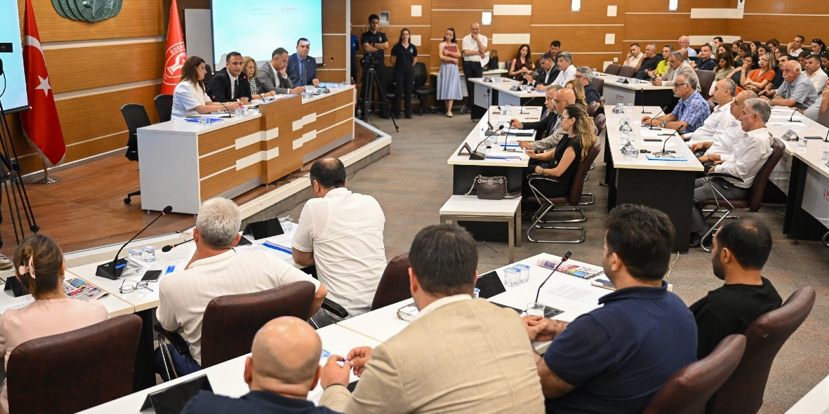 Belediye meclis salonunda düzenlenen toplantıda, protokol masasında oturan yetkililer ve salonu dolduran katılımcılar yer alıyor. Salonun ön kısmında Bornova Belediyesi logosunun bulunduğu bir kürsü ve arkada projeksiyon ekranı bulunurken, katılımcılar U düzenindeki masalarda oturarak sunumu takip ediyor. Salonun sol köşesinde Türk bayrağı yer alıyor.