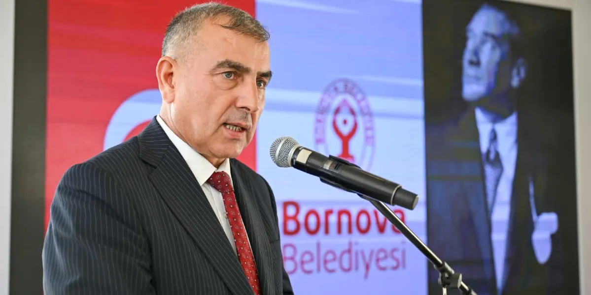 Koyu renkli çizgili bir takım elbise ve kırmızı desenli bir kravat giymiş orta yaşlı bir erkek, önündeki mikrofona konuşma yapıyor. Arka planda Bornova Belediyesi logosu ve Mustafa Kemal Atatürk'ün silüetinin yer aldığı büyük bir ekran bulunuyor.