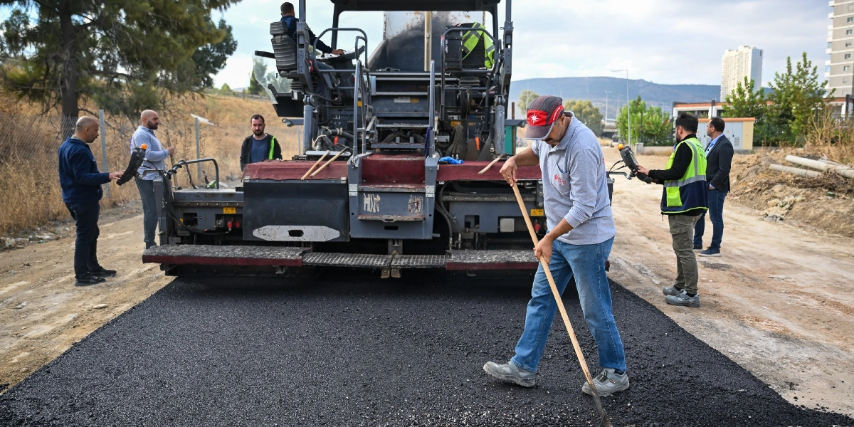 Bornova'da bir yol yapım çalışması sırasında, asfalt serme makinesinin önünde elindeki tırmıkla asfaltı düzelten bir işçi ve onu izleyen diğer belediye çalışanları görülüyor. Arka planda açık bir arazi ve uzakta yüksek binalar yer alıyor.