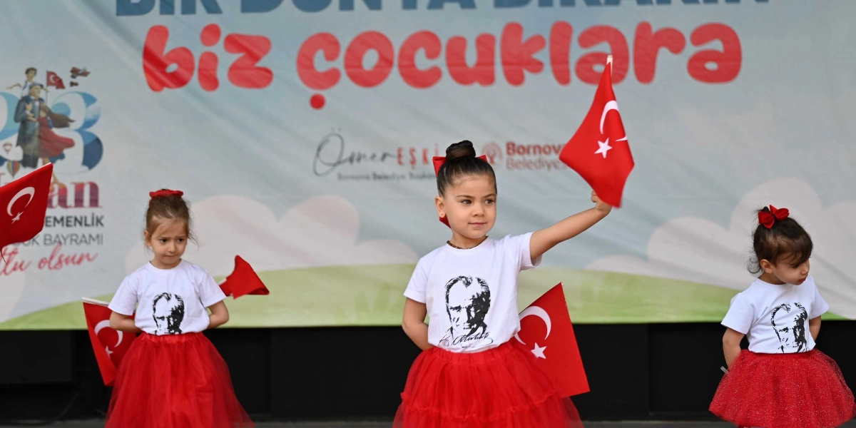 Üzerinde Atatürk baskılı beyaz tişört ve kırmızı tül etek giyen üç küçük kız çocuğu, ellerinde Türk bayraklarıyla 23 Nisan gösterisi için sahnede duruyor. Arka planda 