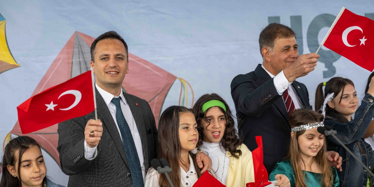 Takım elbiseli iki yetişkin erkek, bir grup çocukla birlikte ellerinde Türk bayrakları tutarak gülümsüyor. Arka planda uçurtma çizimleri olan açık renkli bir fon bulunuyor. Çocuklardan biri başında parlak bir taç takıyor, diğerleri ise neşeli bir ifadeyle etkinliğe katılıyor.