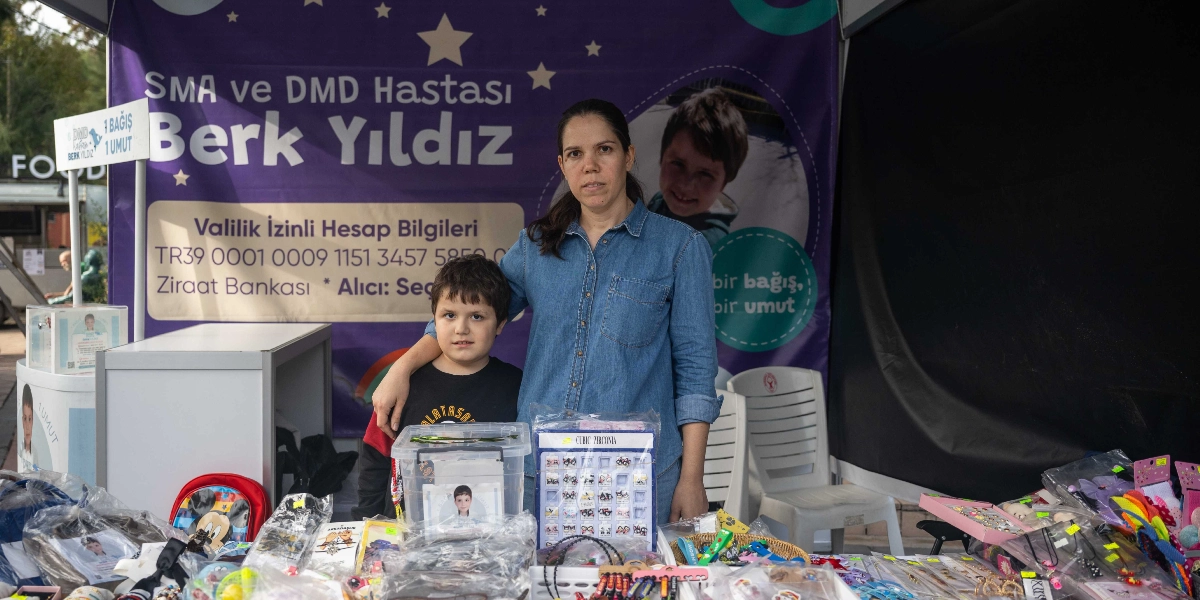 Bir kadın ve yanında duran küçük bir erkek çocuğu, SMA ve DMD hastası Berk Yıldız için açılan bir yardım standının önünde duruyor. Arka planda üzerinde Berk Yıldız'ın fotoğrafının, valilik izinli hesap bilgilerinin ve 
