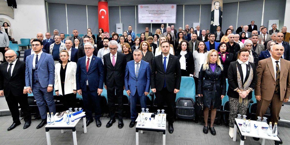 Konferans salonunda bir araya gelmiş, resmi kıyafetli kalabalık bir grup insan ayakta durarak poz veriyor. Arka planda Türk bayrağı, Atatürk portresi ve üzerinde 