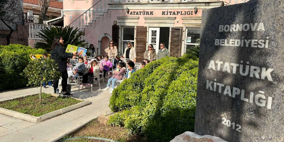 Bornova Belediyesi Atatürk Kitaplığı’nın bahçesinde, bir eğitmen çocuklara kitap okurken, arkada yetişkinler ve çocuklar sandalyelerde oturarak etkinliği izliyor. Ön planda üzerinde Bornova Belediyesi Atatürk Kitaplığı 2012 yazılı büyük bir taş tabela yer alıyor.