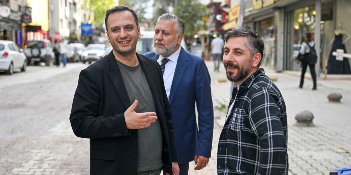 Süvari Caddesi'ndeki yenileme çalışmalarını inceleyen üç erkek, caddenin ortasında sohbet ediyor. Önde siyah ceketli ve gri tişörtlü bir adam gülümseyerek konuşurken, yanında lacivert takım elbiseli, gri sakallı bir kişi ve sağ tarafta ekose gömlekli, sakallı başka bir adam yer alıyor. Arka planda ise dükkanların bulunduğu cadde ve park halindeki araçlar görülüyor.