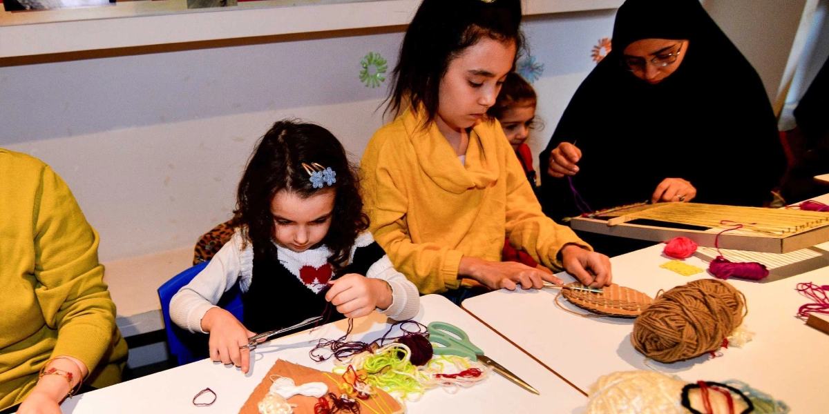 Bir atölye ortamında, beyaz masaların etrafında oturan çocuklar ve bir yetişkin el işi çalışmaları yapıyor. Ön planda, üzerinde kalp deseni bulunan siyah bir yelek giymiş küçük bir kız çocuğu elindeki makasla iplikleri kesiyor. Yanında sarı kazaklı bir kız çocuğu elindeki küçük bir dokuma tezgahıyla çalışırken, arka planda başörtülü bir kadın da benzer bir dokuma işlemiyle uğraşıyor. Masanın üzerinde çeşitli renklerde yün yumakları, makaslar ve küçük dokuma parçaları bulunuyor.