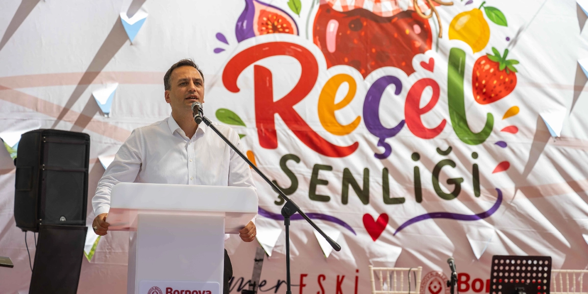 Beyaz gömlekli bir erkek, üzerinde renkli meyve çizimleri ve büyük harflerle Reçel Şenliği yazan bir pankartın önünde, kürsüde mikrofonla konuşma yapıyor.