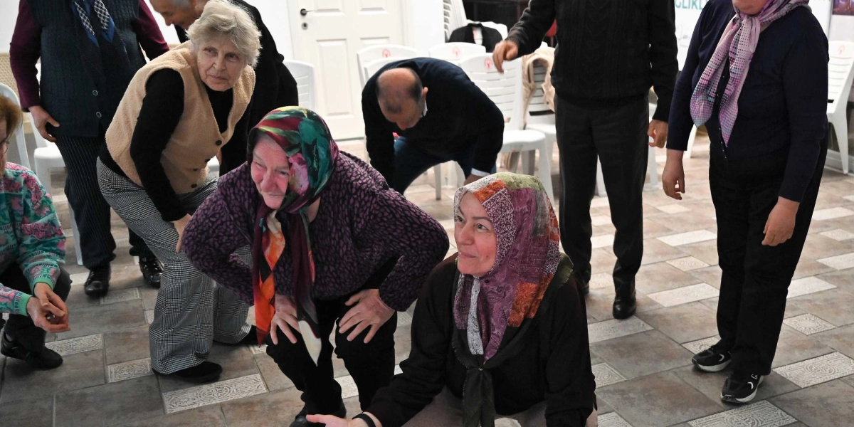 Kapalı bir mekanda yaratıcı drama çalışması yapan bir grup yetişkin, ellerini dizlerine koyarak hafifçe eğilmiş veya çömelmiş pozisyonda duruyor. Katılımcıların bir kısmı başörtülü, bir kısmı ise günlük kıyafetler içinde; hepsi dikkatle bir noktaya odaklanmış durumda. Arka planda beyaz plastik sandalyeler ve zeminde desenli fayanslar görülüyor.