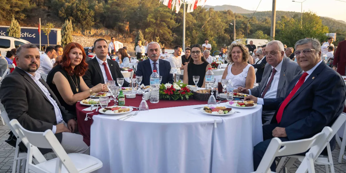 Açık havada düzenlenen bir yemek davetinde, beyaz örtülü yuvarlak bir masanın etrafında oturan, aralarında Bornova Belediye Başkanı Ömer Eşki'nin de bulunduğu bir grup resmi giyimli kişi kameraya bakıyor. Masanın ortasında çiçek aranjmanı, su şişeleri ve tabaklarda yemekler yer alırken, arka planda ağaçlıklı bir alan ve asılı Türk bayrakları görülüyor.