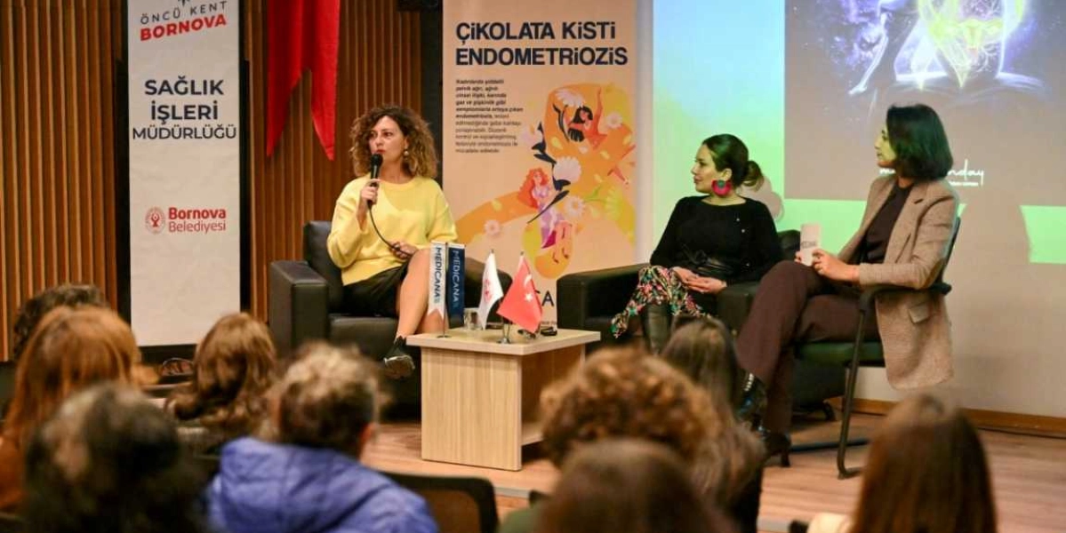 Bornova Belediyesi Sağlık İşleri Müdürlüğü tarafından düzenlenen Endometriozis farkındalık etkinliğinde, üç kadın konuşmacı sahnede siyah koltuklarda oturarak katılımcılara hitap ediyor. Soldaki konuşmacı elinde mikrofonla konuşurken, diğer iki konuşmacı onu dinliyor. Konuşmacıların arkasında üzerinde 