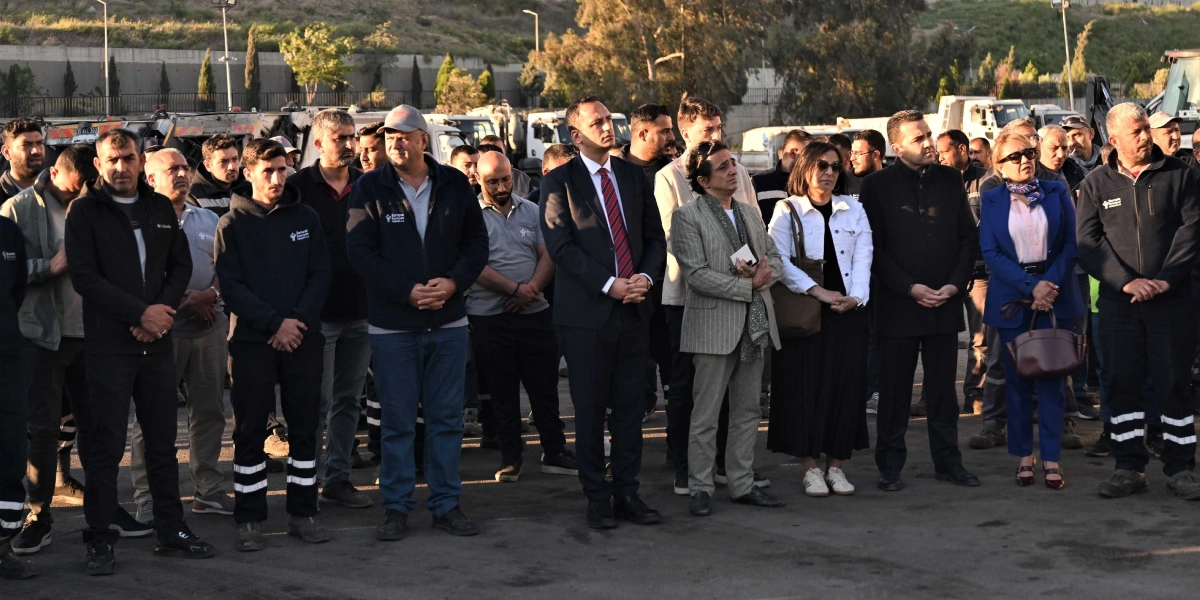 Açık havada, asfalt bir alanda yan yana duran ve ciddiyetle karşıya bakan kalabalık bir grup insan. Grubun ön sırasında, takım elbiseli bir erkek, yanında çizgili ceketli bir kişi, beyaz ceketli bir kadın ve mavi takım elbiseli bir kadın yer alıyor. Diğer katılımcılar ise genellikle günlük kıyafetler ve bazıları üzerinde Bornova Belediyesi logolu iş kıyafetleri ile duruyor. Arka planda ise park halindeki belediye hizmet araçları ve ağaçlıklı bir yamaç görülüyor.