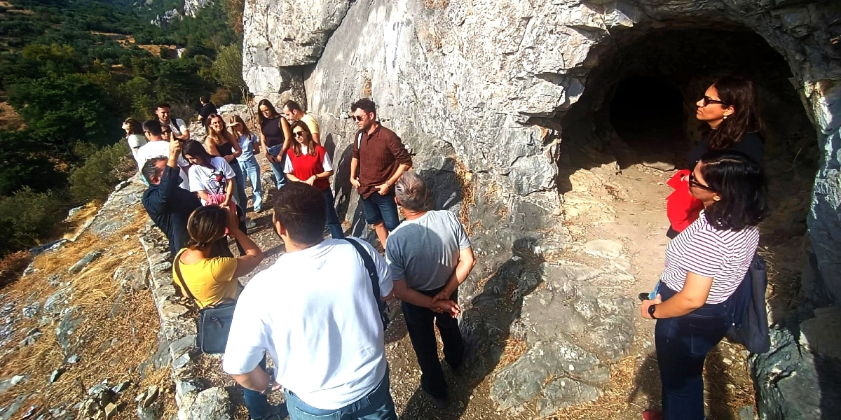 Açık havada, kayalık bir yamaçta bulunan doğal bir mağara girişinin önünde toplanmış bir grup insan. Gruptaki kişiler, rehberlik ettiği anlaşılan birinin anlatımını dinleyerek mağara girişini inceliyor. Arka planda ağaçlarla kaplı bir vadi manzarası görülüyor.