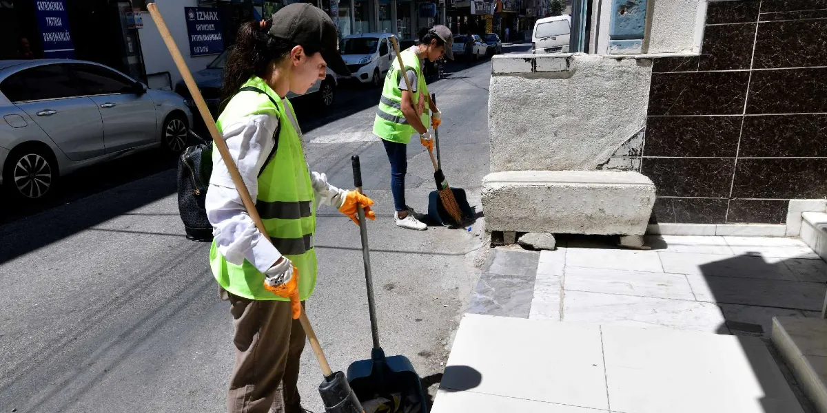Üzerlerinde fosforlu sarı iş yelekleri, şapkalar ve turuncu eldivenler bulunan iki belediye çalışanı, Bornova'da bir sokak kenarındaki kaldırımı ve yolu süpürge ve faraş yardımıyla temizliyor.