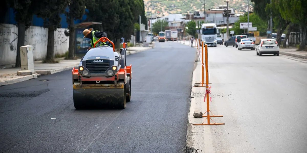 Pınar Caddesi'nde devam eden yol yenileme çalışmaları kapsamında, üzerinde operatörün bulunduğu turuncu ve gri renkli bir silindir aracı yeni dökülen asfaltı düzleştiriyor. Çalışma alanı, trafiğin aktığı şeritten turuncu renkli güvenlik direkleri ve şeritlerle ayrılmış durumda.