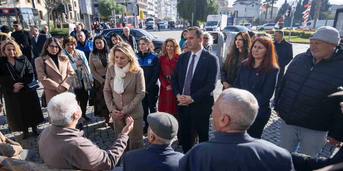 CHP Kadın Kolları Başkanı Asu Kaya ve Bornova Belediye Başkanı Ömer Eşki'nin de aralarında bulunduğu bir grup, açık havada vatandaşlarla bir araya gelerek sohbet ediyor. Asu Kaya bej rengi bir takım elbise ve beyaz bir şal ile ayakta dururken, Ömer Eşki lacivert bir takım elbise ve kravatla yanında yer alıyor. Çevredeki diğer katılımcılar ve vatandaşlar dikkatle konuşmaları dinliyor. Arka planda ise şehir içi trafik ve park halindeki araçların bulunduğu bir cadde görülüyor.