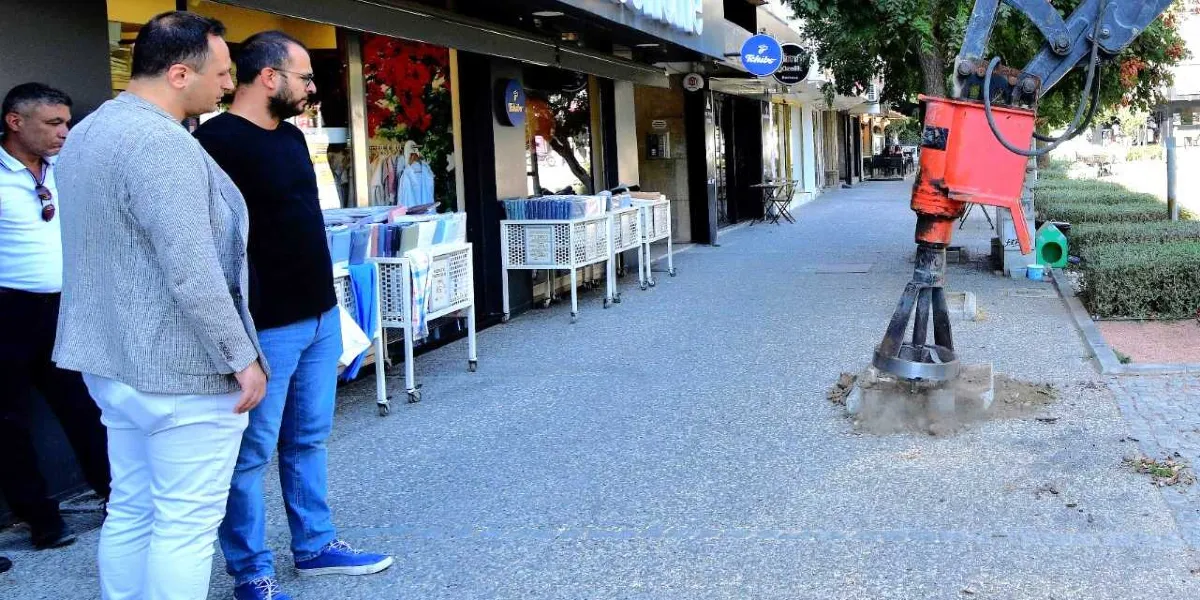 Üç erkek, bir dükkanın önündeki kaldırımda durmuş, bir iş makinesinin açtığı çukuru izliyor. Turuncu renkli bir hidrolik delici, kaldırımın bir kısmını kazarak yeni bir ağaç dikimi için hazırlık yapıyor. Dükkanın önünde sergilenen ürün stantları ve arka planda ağaçlıklı bir yol görülüyor.