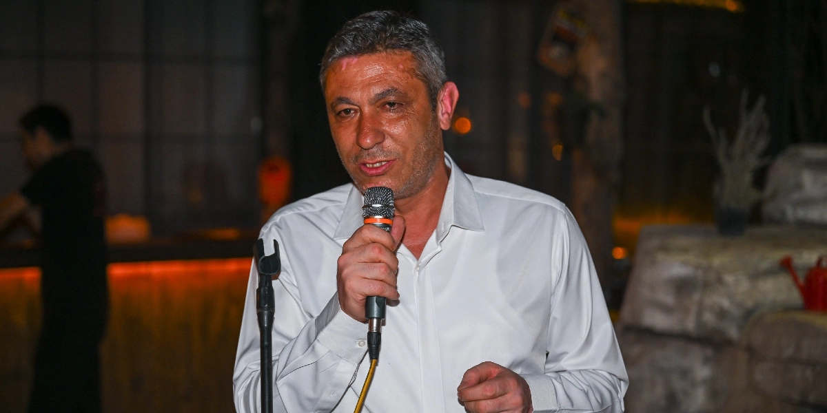 Beyaz gömlekli, kır saçlı bir erkek, iç mekanda elinde mikrofon tutarak konuşma yapıyor.