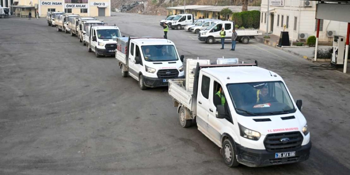 Bornova Belediyesi'ne ait beyaz renkli kamyonetler, belediye yerleşkesindeki geniş bir alanda tek sıra halinde dizilmiş bekliyor. Araçların üzerinde T.C. Bornova Belediyesi yazısı ve logoları bulunuyor. Arka plandaki binanın üzerinde 