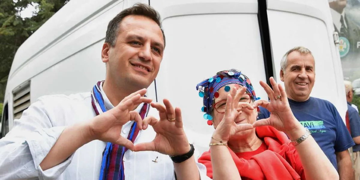Beyaz gömlekli ve boynunda yöresel desenli bir şal olan Başkan Filiz Ceritoğlu Sengel ile başında renkli pullarla süslü bir başlık bulunan bir kadın, elleriyle kalp işareti yaparak gülümsüyor. Arka planda beyaz bir minibüs ve üzerinde mavi tişörtüyle gülümseyen bir erkek yer alıyor.