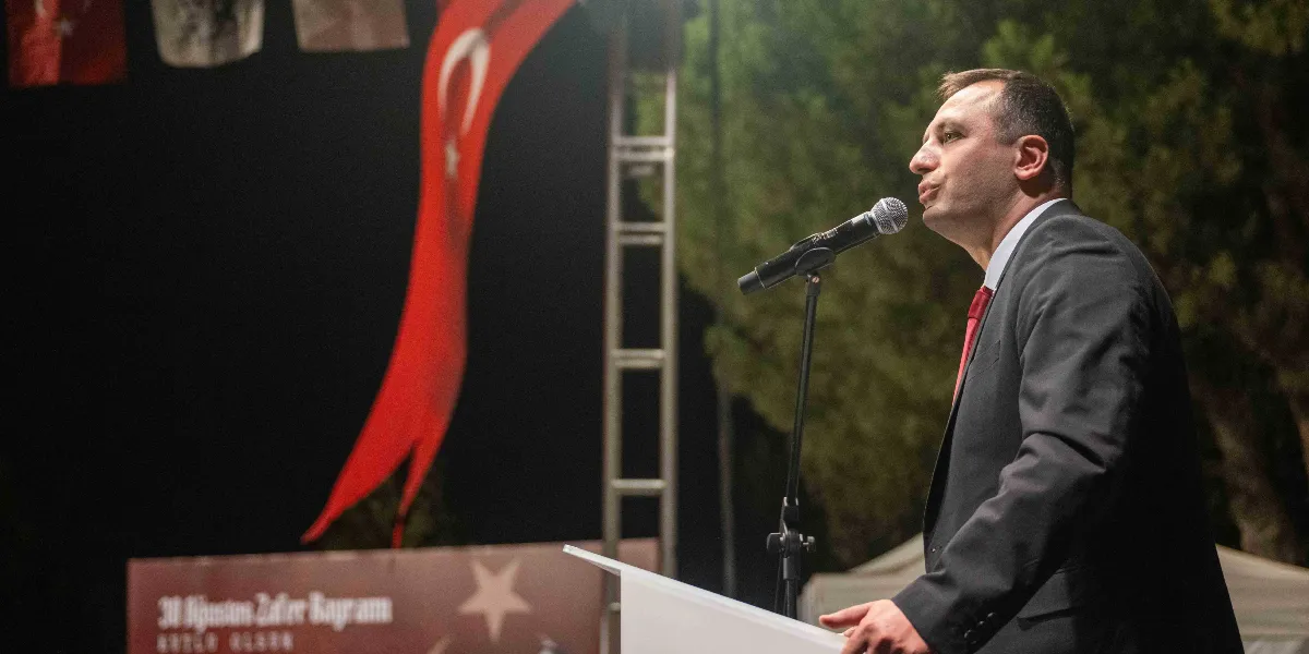 Koyu renkli takım elbise ve kırmızı kravat giymiş bir erkek, gece vakti açık havada düzenlenen bir etkinlikte kürsüde mikrofon başında konuşma yapıyor. Arka planda dev bir Türk bayrağı asılı duruyor ve üzerinde 