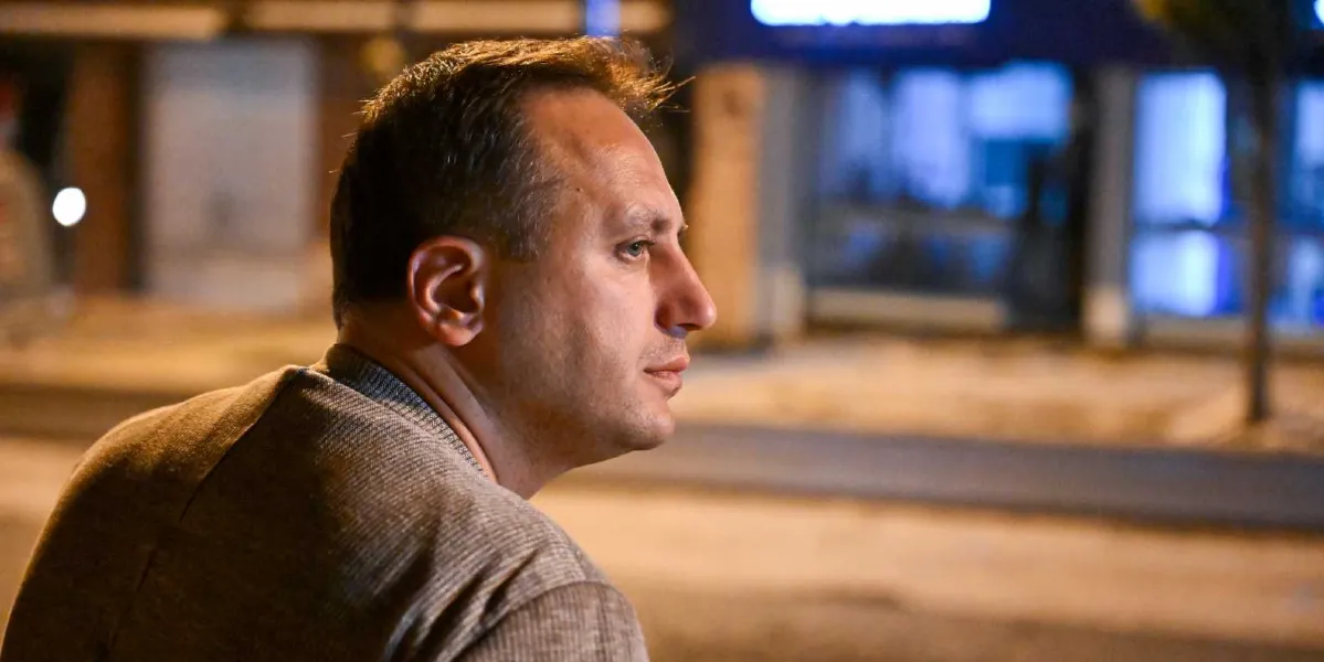 Gece vakti dışarıda, gri bir kazak giymiş orta yaşlı bir adam profilden sağ tarafa doğru bakıyor. Arka planda bulanık bir şekilde şehir caddesi ve ışıklar görülüyor.