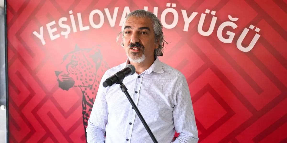 Kır saçlı, sakallı ve açık renkli düğmeli bir gömlek giyen bir erkek, önündeki mikrofona konuşma yapıyor. Arka planda üzerinde 