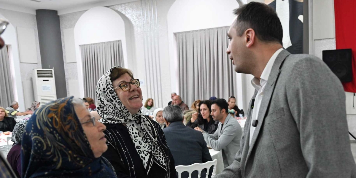 Gri ceketli bir erkek, dayanışma yemeğinde başörtülü iki kadınla güler yüzle sohbet ediyor. Arka planda ise masalarda oturan diğer katılımcılar ve beyaz perdelerle dekore edilmiş geniş bir salon görülüyor.