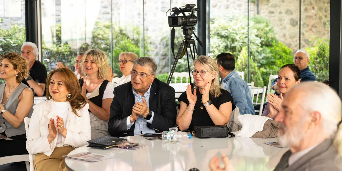 Modern bir toplantı salonunda, beyaz yuvarlak bir masanın etrafında oturan ve alkışlayan bir grup insan. Ön planda, beyaz ceketli bir kadın, lacivert ceketli bir erkek ve siyah bluzlu bir kadın dikkatle izleyip alkışlarken görülüyor. Arka planda ise bir tripod üzerinde kurulu profesyonel bir kamera ve cam bölmelerin ardında yeşil bir bahçe manzarası yer alıyor. Katılımcıların çoğu gülümseyerek bir sunumu veya konuşmayı takip ediyor.