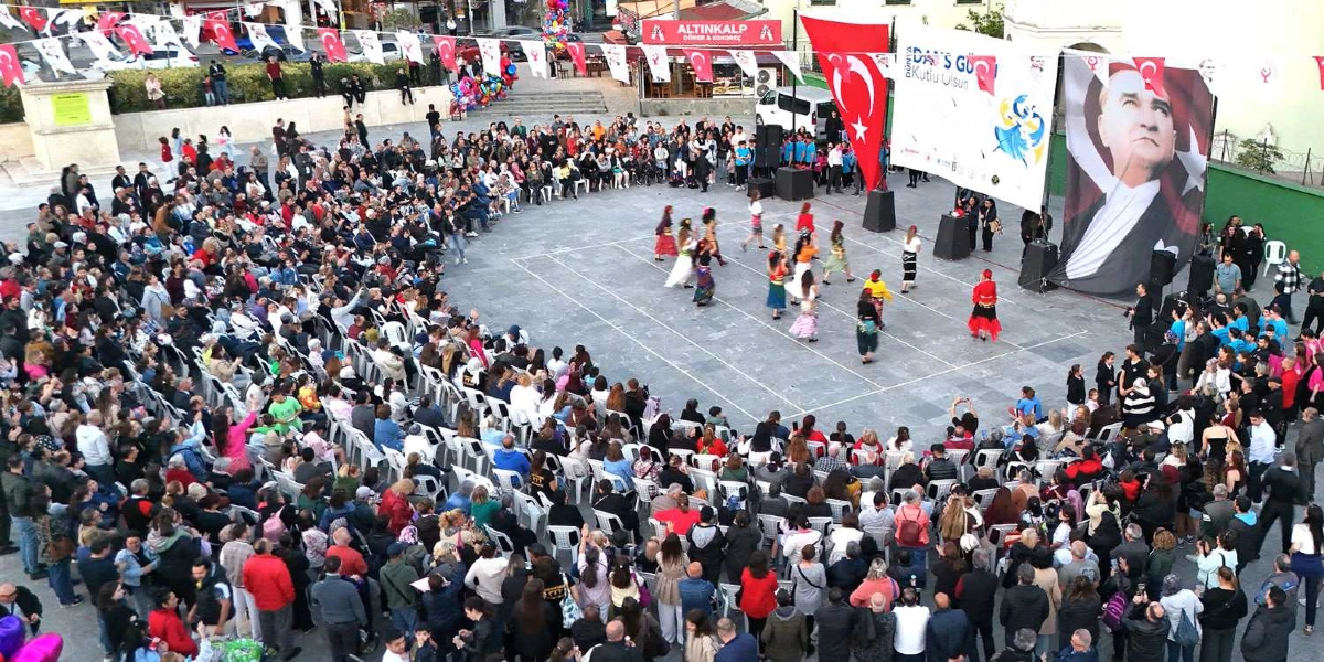 Bornova'da açık bir meydanda düzenlenen Dünya Dans Günü kutlamasında, renkli kostümler giymiş bir grup dansçı orta alanda performans sergiliyor. Meydanın çevresine dizilmiş çok sayıda izleyici, beyaz plastik sandalyelerde oturarak gösteriyi takip ediyor. Alanın bir köşesinde üzerinde 