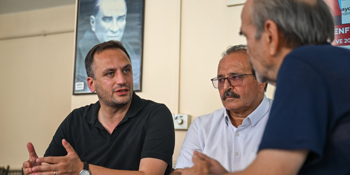 Başkan Eşki, kapalı bir mekanda iki vatandaşla karşılıklı oturarak sohbet ediyor. Başkan siyah polo yaka tişört giymiş, ellerini kullanarak konuşuyor. Yanında gözlüklü ve bıyıklı, beyaz gömlekli bir beyefendi ve sırtı kameraya dönük başka bir kişi daha bulunuyor. Arka plandaki duvarda Mustafa Kemal Atatürk'ün çerçeveli bir portresi asılı.