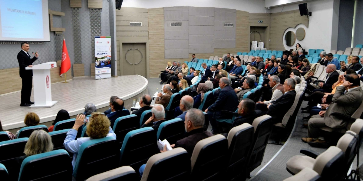 Modern bir konferans salonunda, kürsüde konuşma yapan bir yetkili ve onu dinleyen kalabalık bir grup muhtar yer alıyor. Salonun ön tarafında, üzerinde 