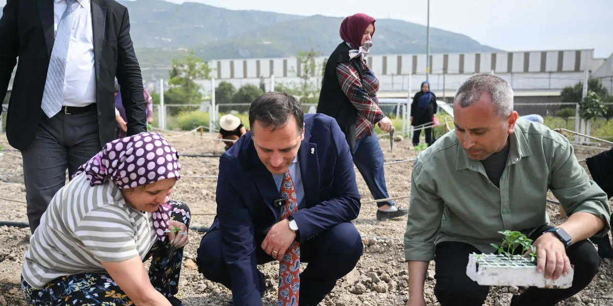 Açık havada, toprak bir alanda bir grup insan fidan dikimi yapıyor. Ön planda, çizgili tişörtlü ve puantiyeli başörtülü bir kadın, lacivert takım elbiseli ve desenli kravatlı bir erkek ile haki renkli gömlek giyen başka bir erkek toprağa fidan dikmek için çömelmiş durumda. Arka planda ise elinde bahçe aleti tutan, başörtülü bir kadın ve diğer katılımcılar yer alıyor. Uzakta dağ manzarası ve endüstriyel yapılar görülüyor.