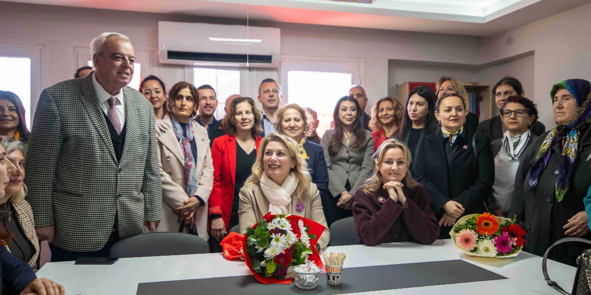 İç mekan bir toplantı odasında, masanın etrafında bir araya gelmiş kalabalık bir grup insan yer alıyor. Masanın ön tarafında oturan CHP Kadın Kolları Başkanı Asu Kaya, beyaz şalı ve bej ceketiyle dikkat çekerken, yanında bordo hırkasıyla oturan bir kadın bulunuyor. Masanın üzerinde iki adet çiçek buketi ve bir bardak kürdan yer alıyor. Grubun sol tarafında, ekose ceketli ve kravatlı bir erkek ayakta duruyor. Arka planda ise farklı yaş ve giyim tarzlarında çok sayıda kadın ve erkek, gülümseyerek kameraya bakıyor. Ortamda resmi bir toplantı veya ziyaret atmosferi hakim.
