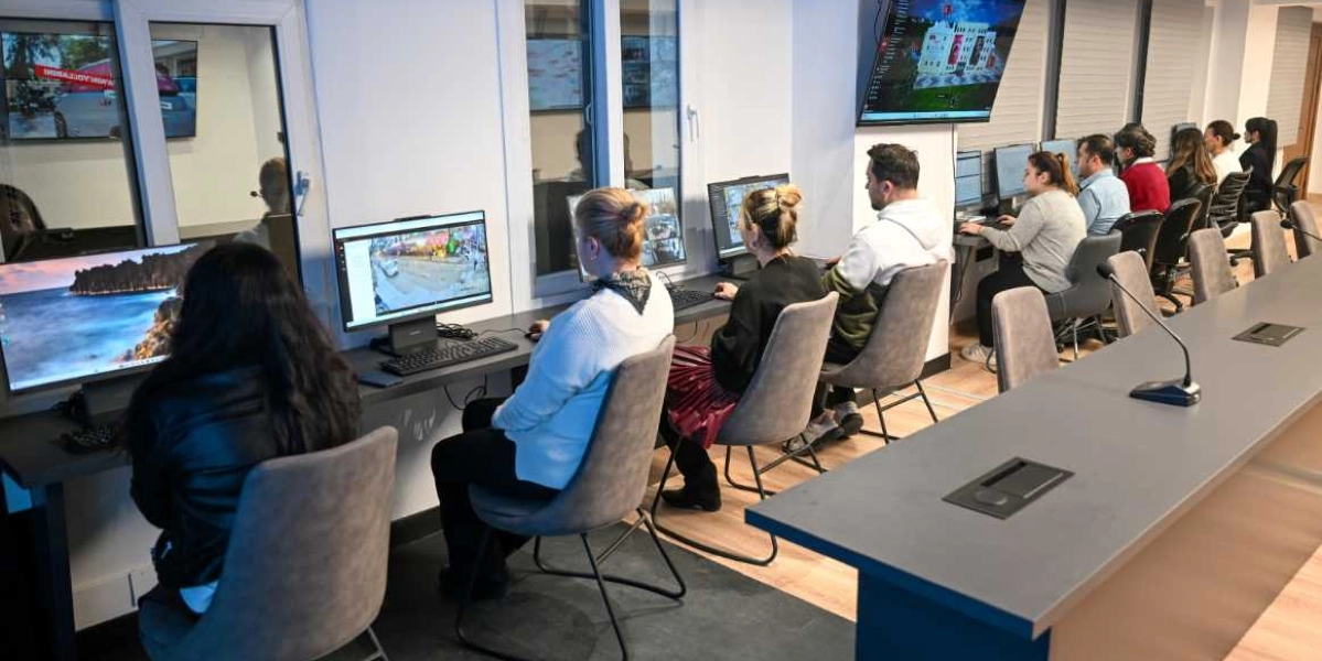 Modern bir ofis ortamında, yan yana dizilmiş bilgisayar masalarında çalışan bir grup insan. Çalışanlar önlerindeki ekranlardan güvenlik kamerası görüntülerini ve dijital haritaları takip ediyor. Arka planda duvara monte edilmiş büyük bir ekranda Bornova Belediyesi binasının üç boyutlu dijital modeli ve Türk bayrağı görünüyor. Odada uzun bir çalışma masası, gri ofis sandalyeleri ve profesyonel bir izleme merkezi atmosferi hakim.