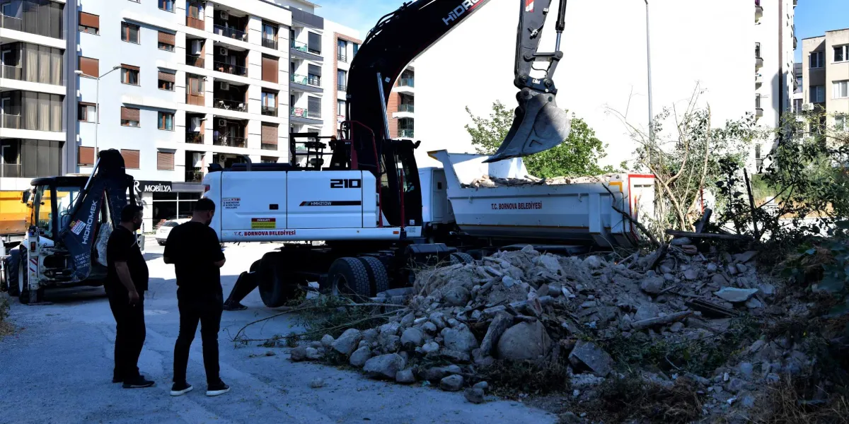 Bornova Belediyesi'ne ait bir iş makinesi, biriken moloz yığınlarını temizleyerek kamyona yüklüyor. Çalışmaları iki kişi yakından takip ederken, arka planda çok katlı konut binaları yer alıyor.