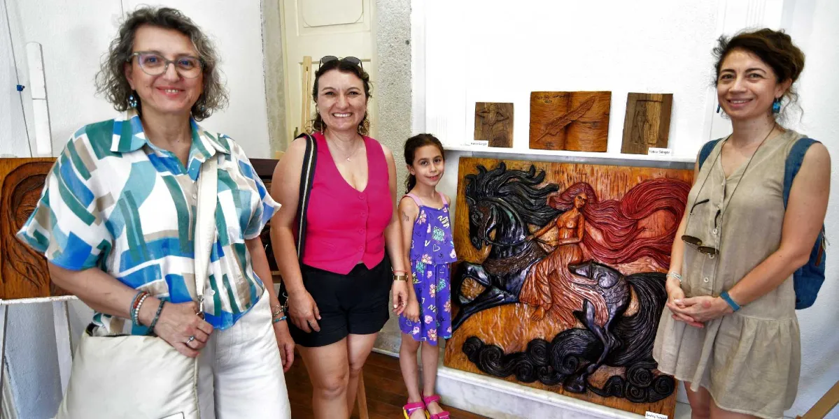 Beyaz duvarlı bir sanat galerisinde, ahşap oyma eserlerin önünde yan yana duran dört kadın ve bir kız çocuğu gülümsüyor. En soldaki kadın mavi, yeşil ve beyaz desenli bir gömlek ve beyaz pantolon giyiyor. Yanındaki kadın pembe bir yelek ve siyah şort, yanındaki küçük kız ise mor desenli bir elbise giyiyor. En sağdaki kadın ise bej rengi kolsuz bir elbise tercih etmiş. Arka planda duvarda sergilenen ahşap oyma eserler arasında, üzerinde bir at ve atın üzerinde uzun saçlı bir kadın figürünün işlendiği büyük, detaylı bir tablo dikkat çekiyor.